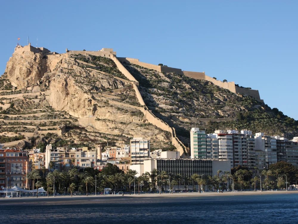 Santa Bárbara Castle's medieval walls and towers rising above Alicante city and the Mediterranean coast, with Postiguet beach glinting in the afternoon sun below