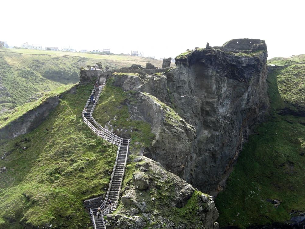 The ruined walls of Tintagel Castle clinging to dramatic Atlantic clifftops in Cornwall, with the steel footbridge spanning the chasm between the island and mainland wards