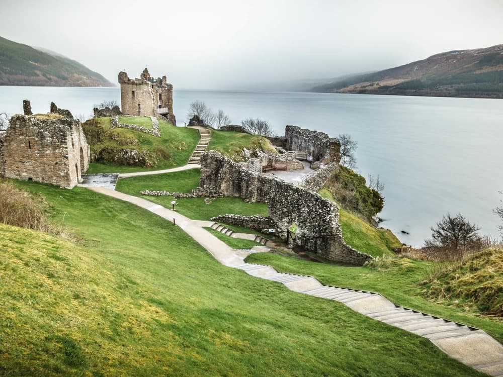 Urquhart Castle's roofless towers and gatehouse ruins on the wooded shore of Loch Ness, with the dark Highland water stretching into the mist behind